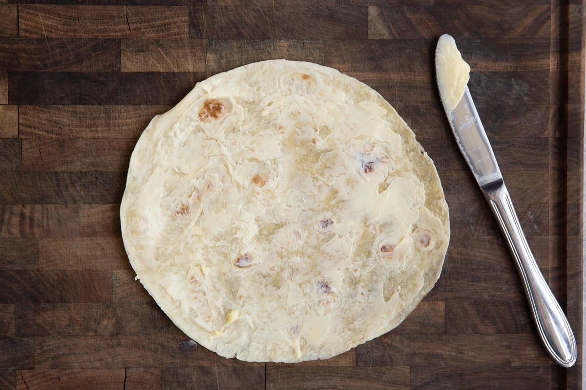 Buttered tortilla on a wooden cutting board with a butter knife with butter on it to the side.