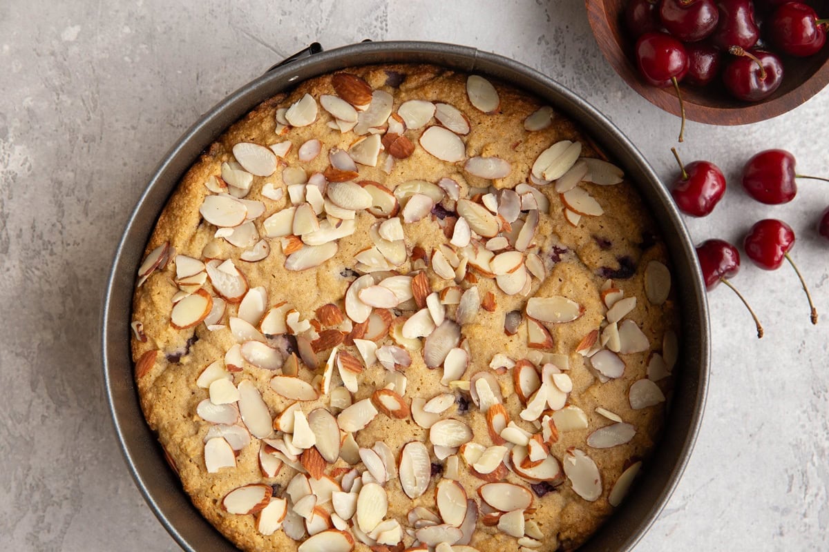 Spring form cake pan with cake in the pan sprinkled with sliced almonds and a bowl of fresh cherries to the side.