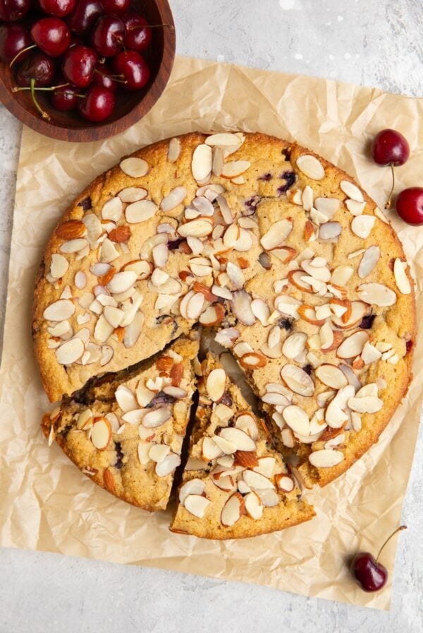 Cherry cake cut into slices on a sheet of parchment paper with fresh cherries scattered around and a small bowl of fresh cherries to the side.