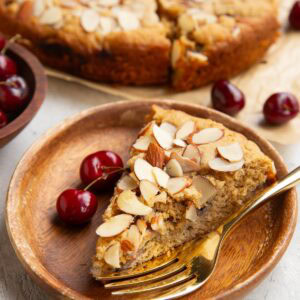 Slice of cherry cake on a wooden plate and two fresh cherries to the side. The rest of the cake in the background.