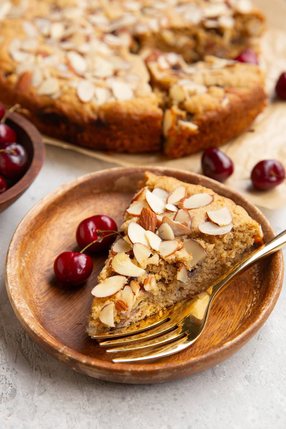 Slice of cherry cake on a wooden plate and two fresh cherries to the side. The rest of the cake in the background.