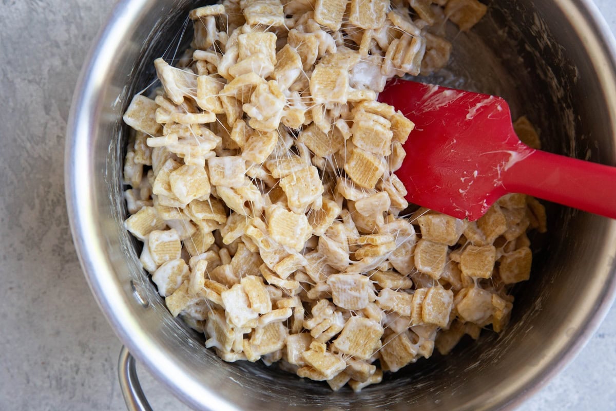 Cereal with melted marshmallows mixed in inside of a saucepan with a red rubber spatula.