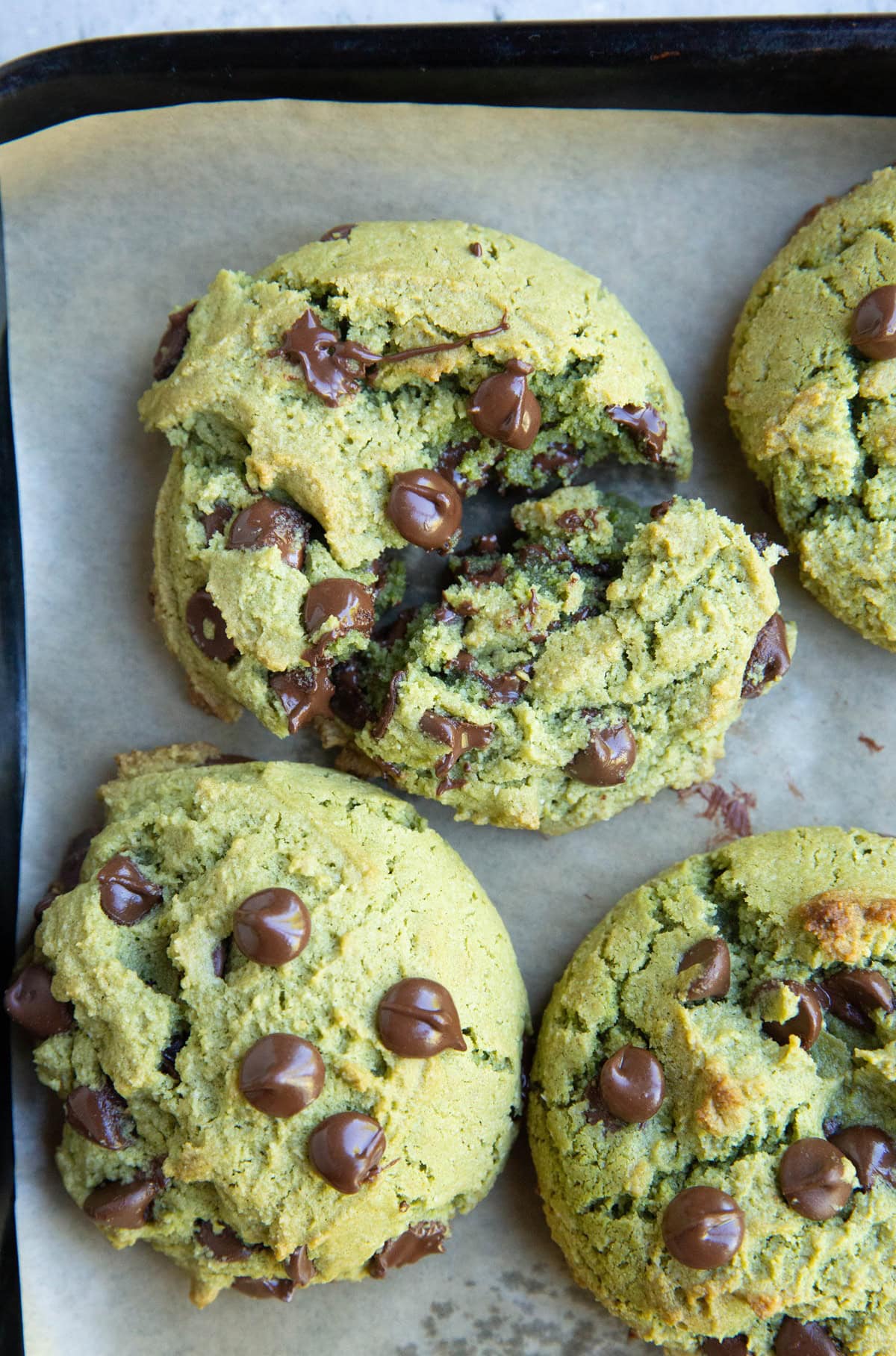 Cookie sheet with green pistachio butter cookies with chocolate chips and one of the cookies is broken in half.
