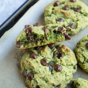 Pistachio cookies on a baking sheet with one of them sliced in half to expose the gooey center.