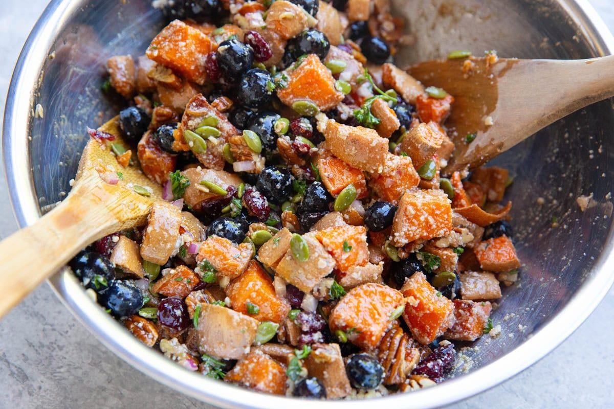 Stainless steel mixing bowl with sweet potato salad inside, mixed up with wooden spoons and ready to serve.