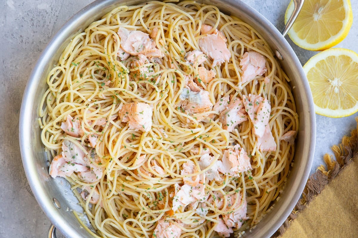 Stainless steel skillet full of salmon pasta with a sliced lemon and a golden napkin to the side.