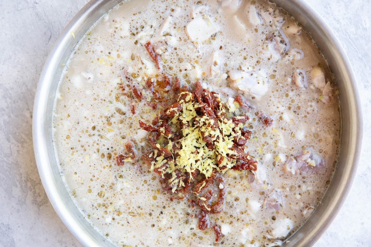 Stainless steel skillet with chicken thighs, orzo, sun-dried tomatoes, lemon zest, broth and coconut milk, ready to be boiled.
