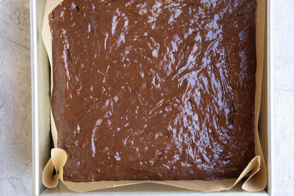 9-inch square baking pan lined in parchment paper with chocolate cake batter inside, ready to go into the oven.