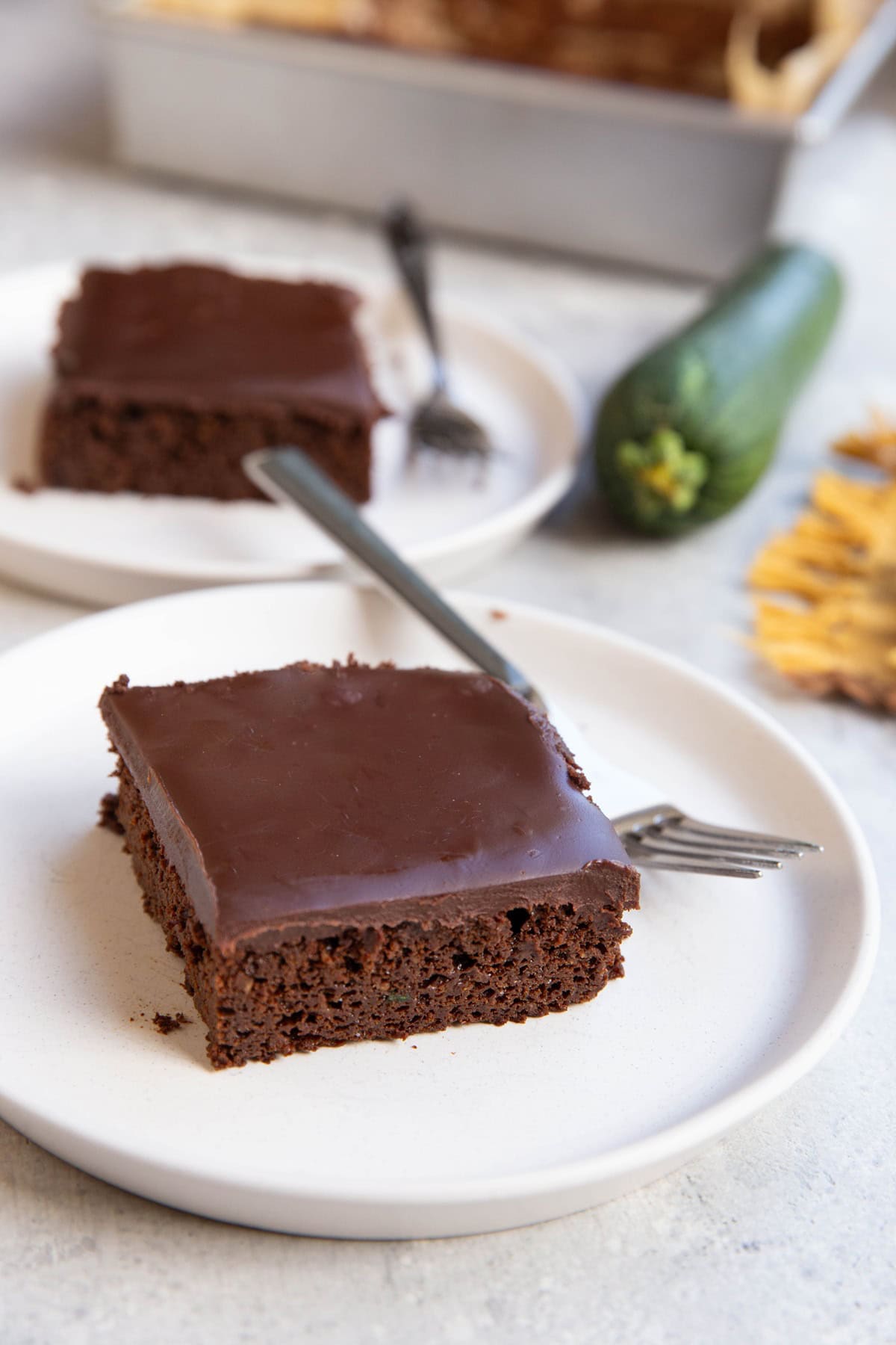 Two slices of chocolate zucchini cake covered frosted with chocolate frosting and a fresh zucchini in the background.