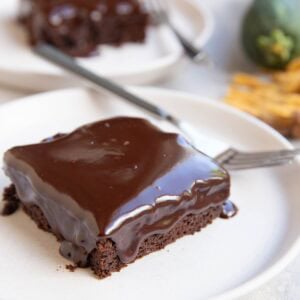 Two slices of zucchini chocolate cake with chocolate ganache melting down the top and sides on white plates and forks, ready to serve. A zucchini squash and a golden napkin in the background.