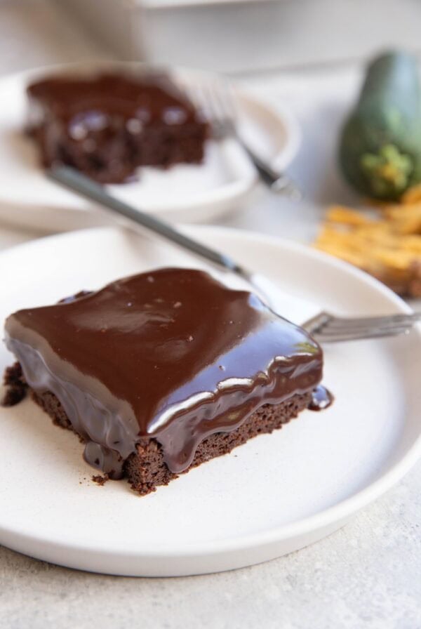 Two slices of zucchini chocolate cake with chocolate ganache melting down the top and sides on white plates and forks, ready to serve. A zucchini squash and a golden napkin in the background.