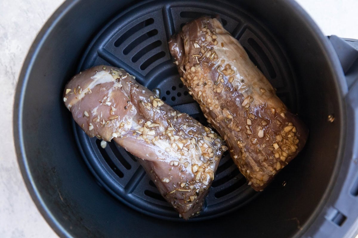 Air fryer basket with two pieces of marinated pork tenderloin inside, ready to cook.
