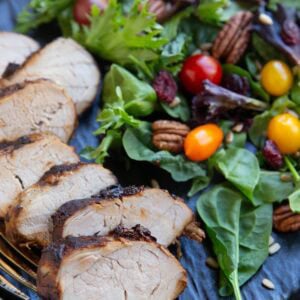 Sliced pork tenderloin on a black plate with a green salad to the side with tomatoes, dried cranberries, sunflower seeds, and pecans.