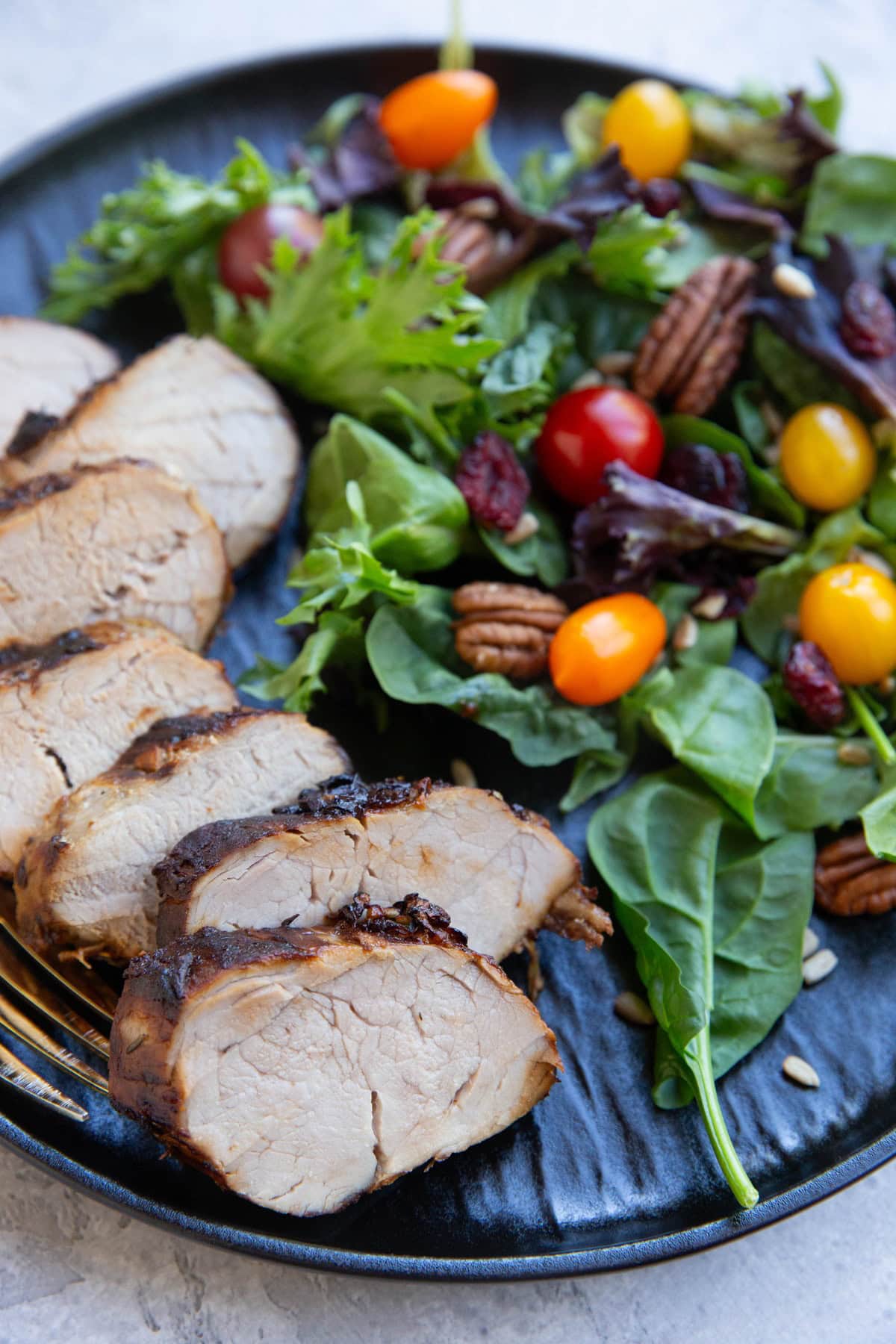 Sliced pork tenderloin on a black plate with a green salad to the side with tomatoes, dried cranberries, sunflower seeds, and pecans.