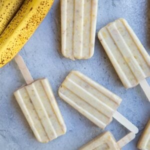 Seven banana popsicles on a blue backdrop with two ripe bananas to the side.