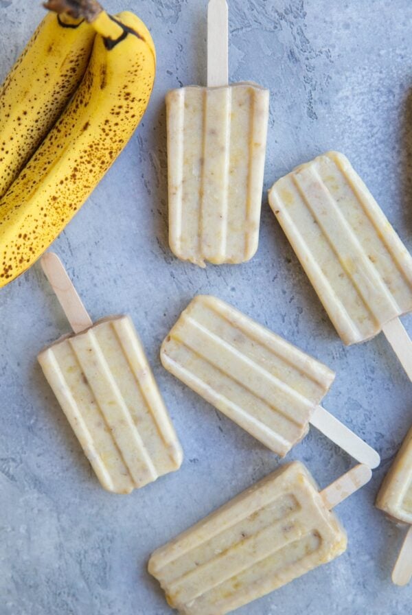 Seven banana popsicles on a blue backdrop with two ripe bananas to the side.