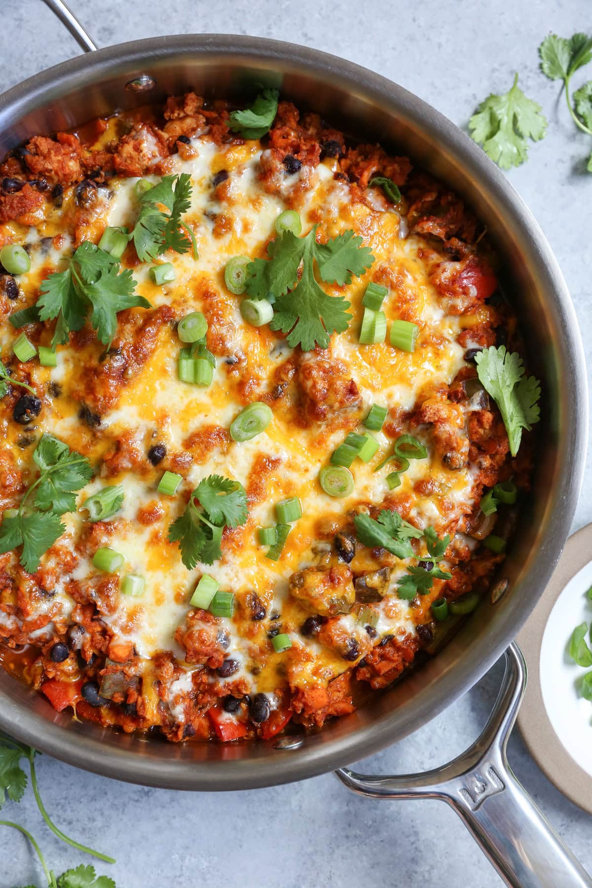 Stainless steel skillet with turkey enchilada with black beans, rice, enchilada sauce, and cheese topped with cilantro and green onions.