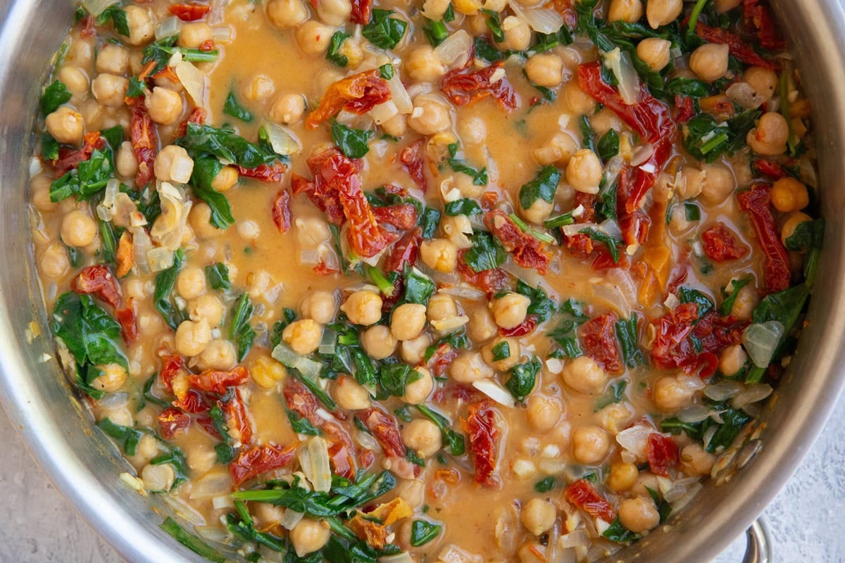 Stainless steel skillet with chickpeas in a creamy sun-dried tomato sauce with spinach.