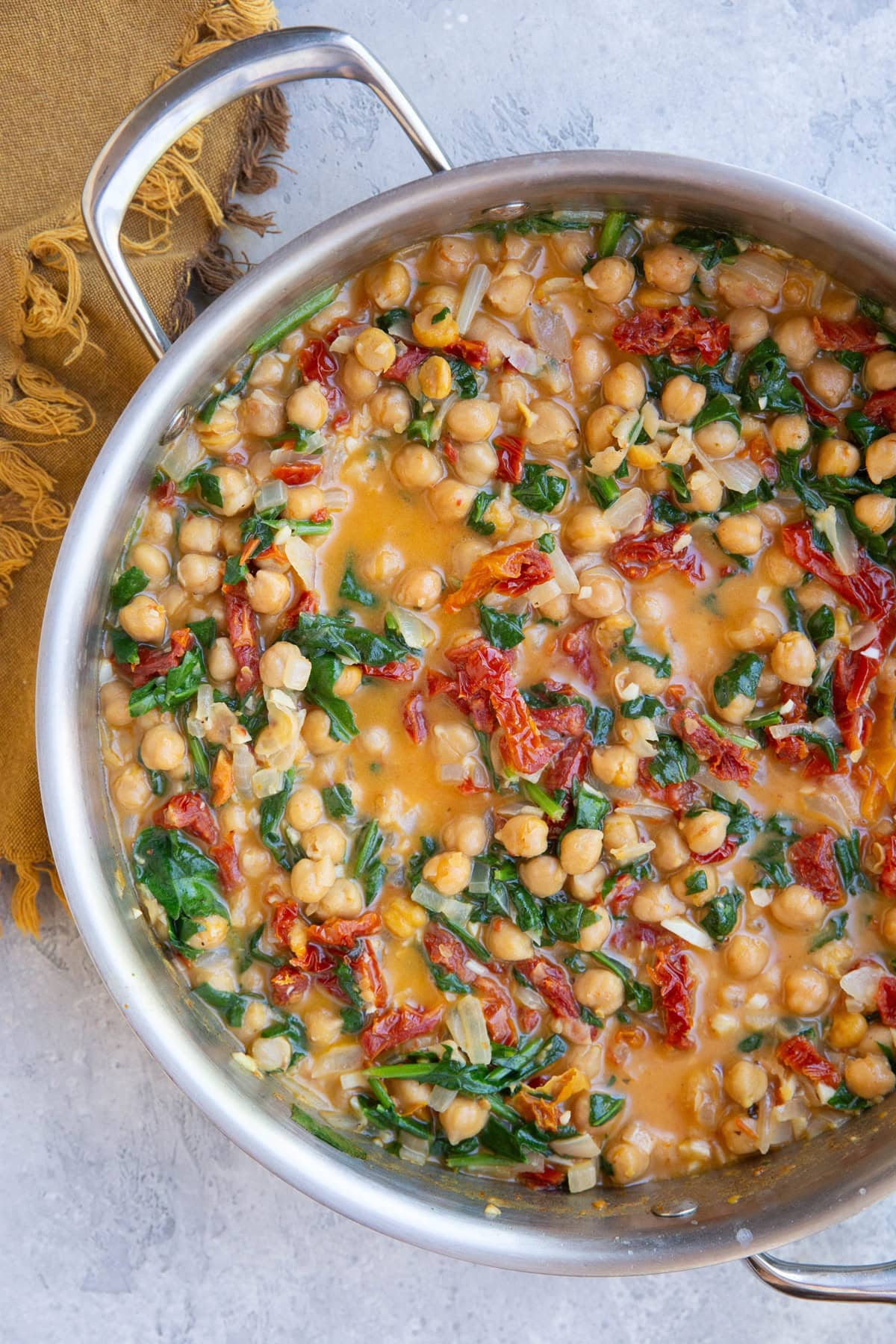 Stainless steel skillet full of sun-dried tomato creamy chickpeas with spinach and onion and a golden napkin to the side.