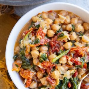 White bowl full of marry me chickpeas with a golden spoon and the skillet with the rest of the meal to the side.