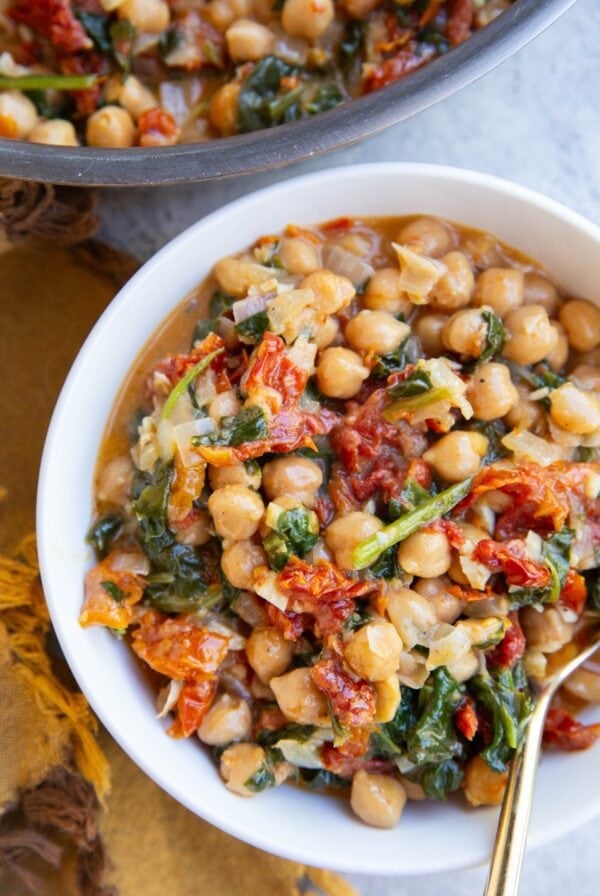 White bowl full of marry me chickpeas with a golden spoon and the skillet with the rest of the meal to the side.