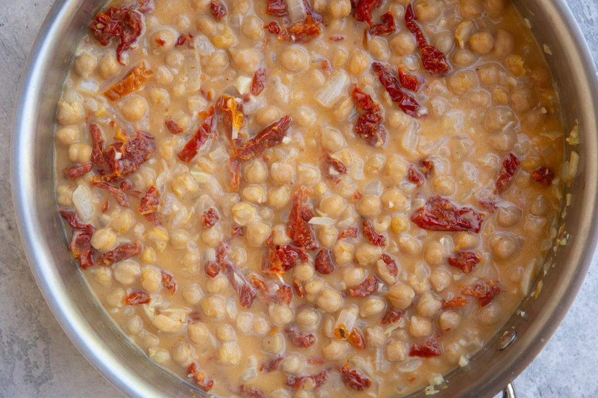 Stainless steel skillet with creamy sauce, chickpeas and sun-dried tomatoes cooking together.