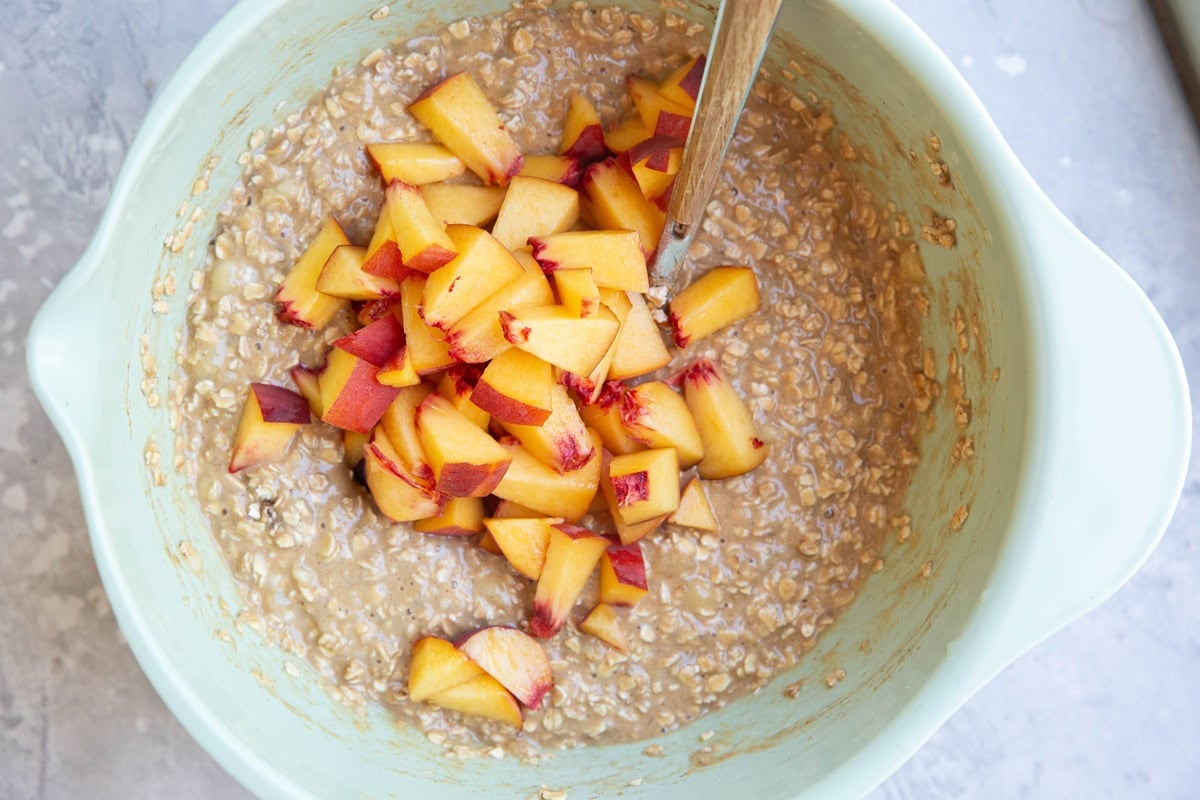 Oatmeal mixture in a mixing bowl with chopped peaches on top, ready to be mixed in.