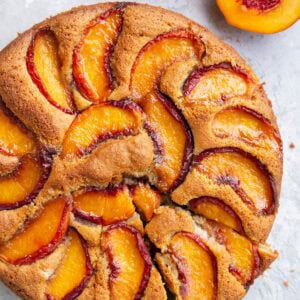 Peach cake one a light blue surface with two slices cut and a fresh peach cut in half to the side.