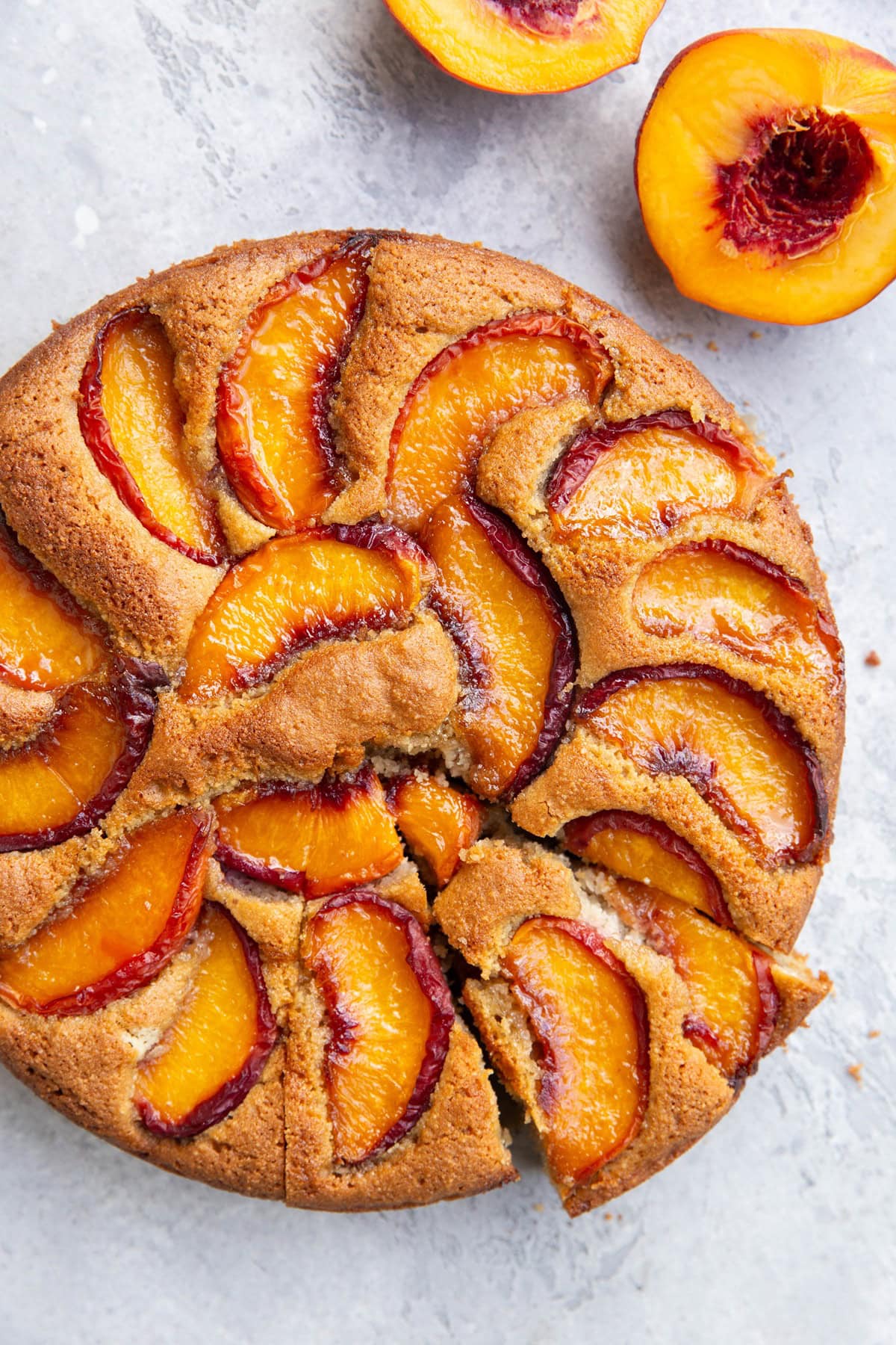 Peach cake one a light blue surface with two slices cut and a fresh peach cut in half to the side.