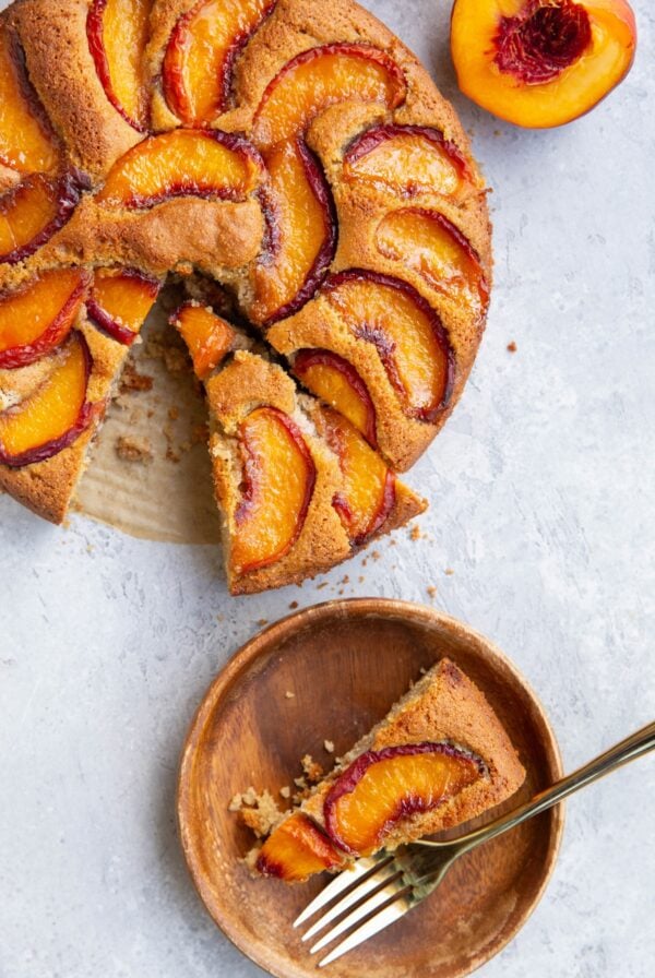 Slice of peach cake on a wooden plate with a golden fork and the rest of the peach cake to the side with another slice cut and a fresh peach half to the side.