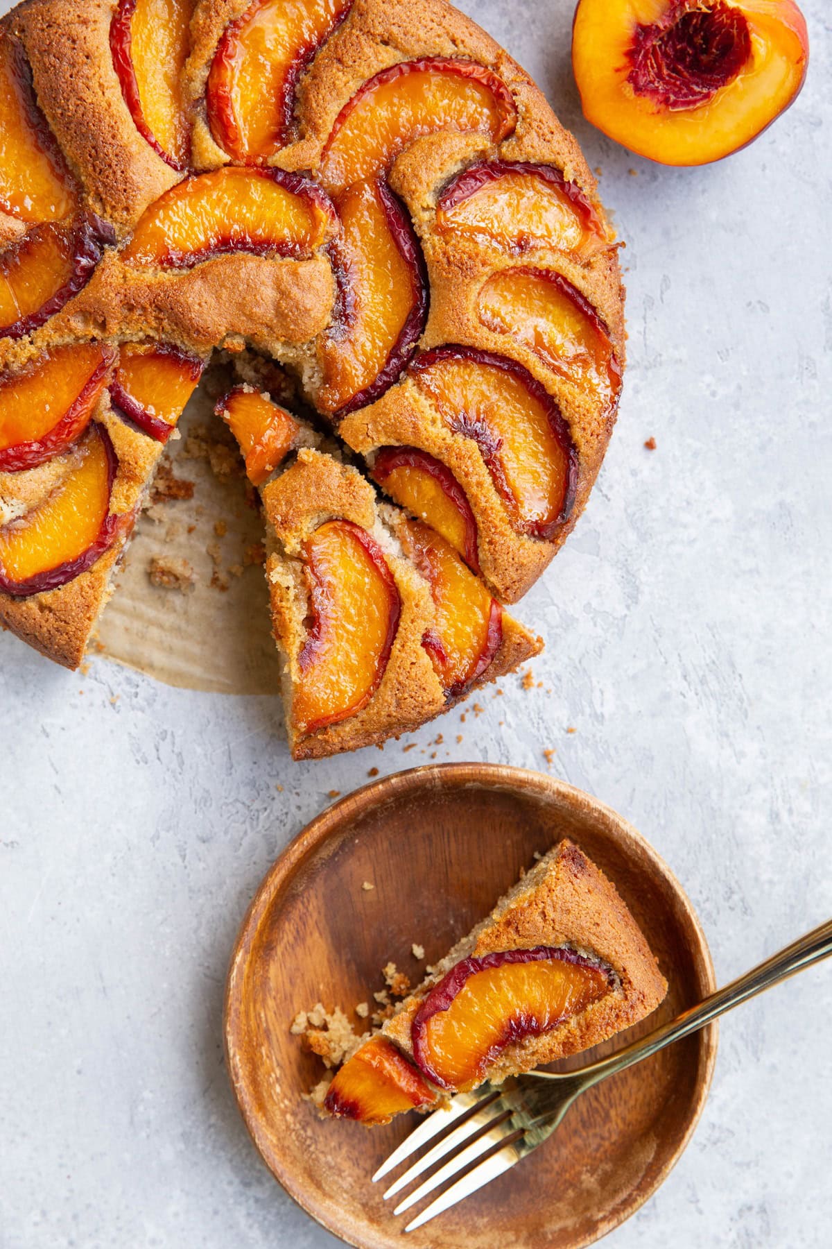 Slice of peach cake on a wooden plate with a golden fork and the rest of the peach cake to the side with another slice cut and a fresh peach half to the side.