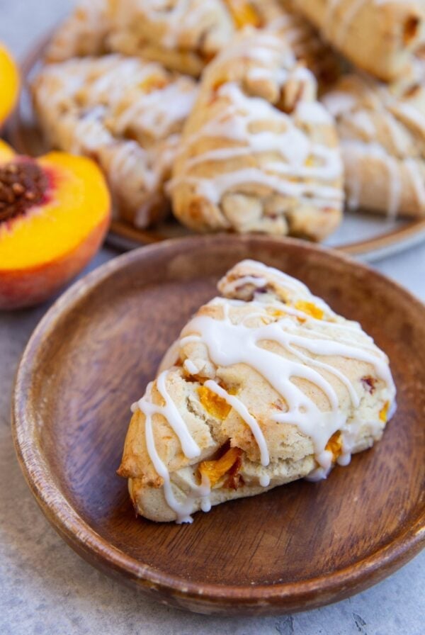 Wooden plate with a peach scone and the rest of the sconed in the background and a fresh peach.