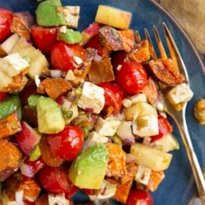 Blue plate full of roasted sweet potatoes, apples, tomatoes, feta, and more with a gold fork to the side for eating.