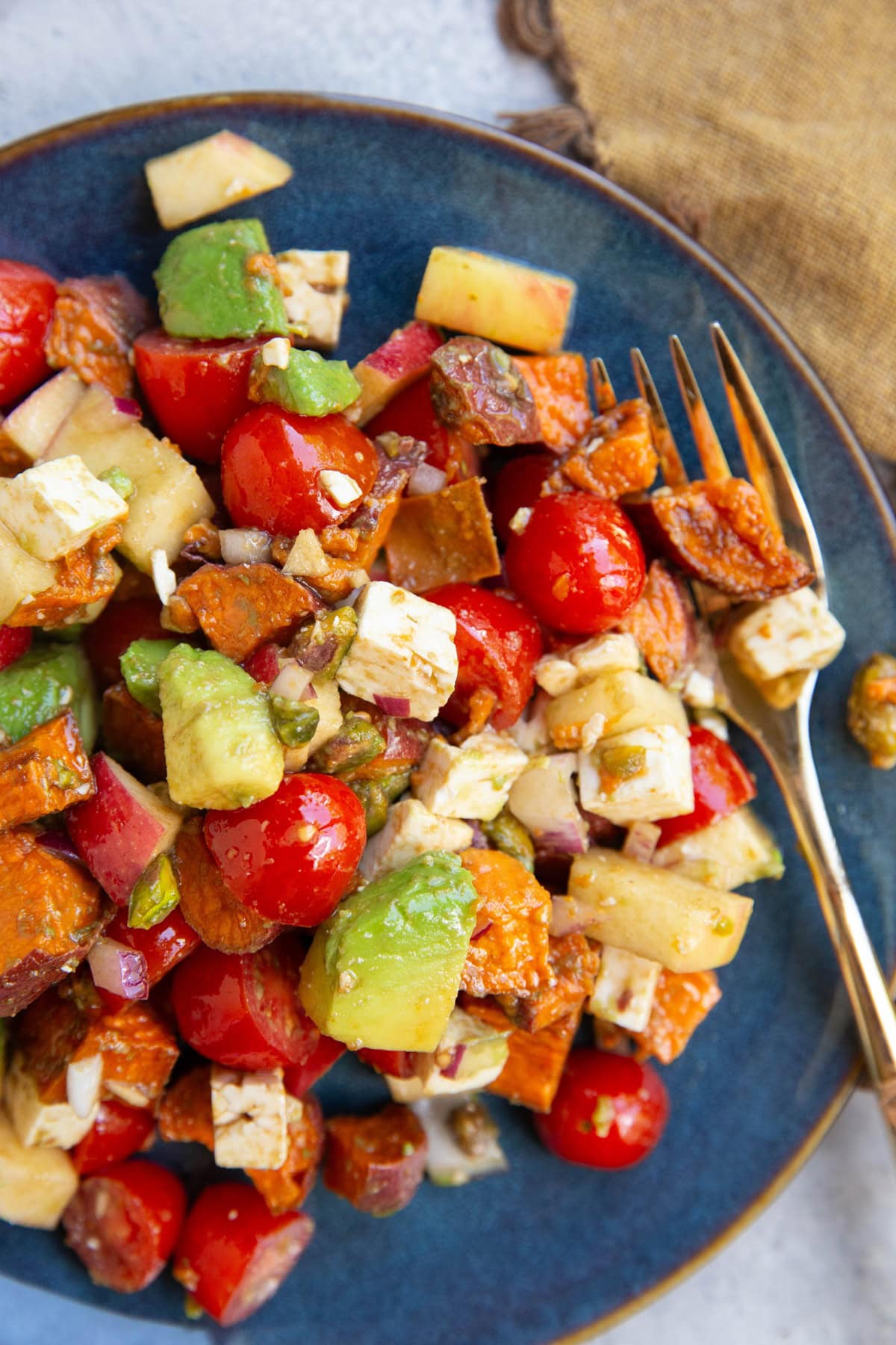 Blue plate full of roasted sweet potatoes, apples, tomatoes, feta, and more with a gold fork to the side for eating.