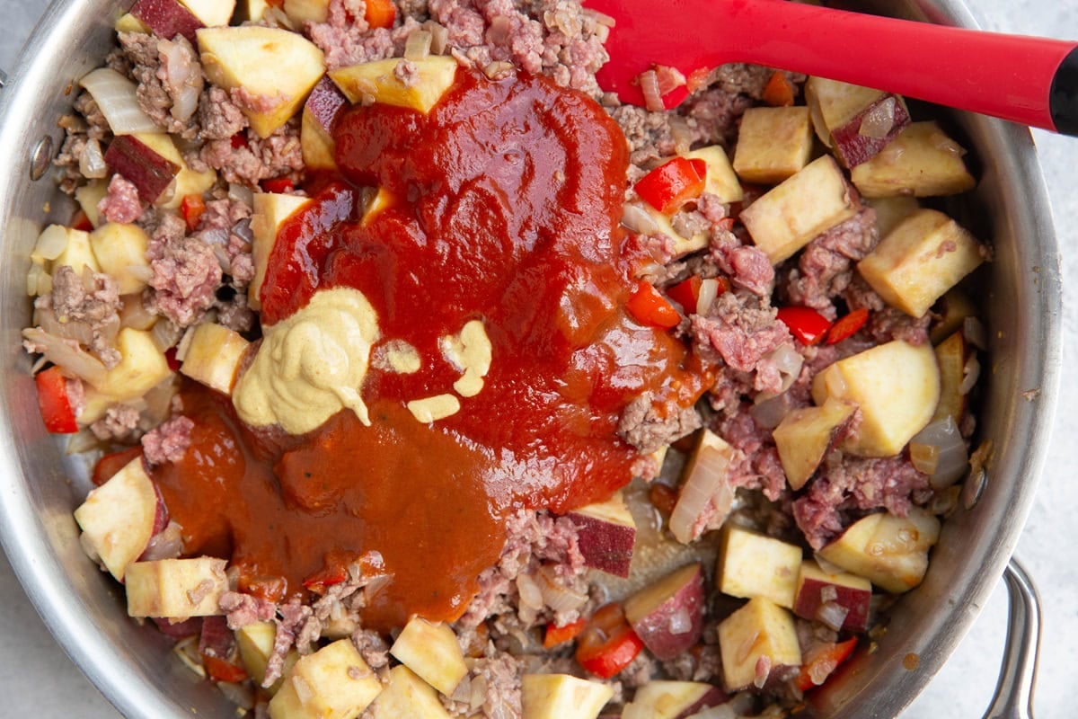 Ground beef, sweet potatoes, onion, bell pepper, ketchup, mustard, bbq sauce in a skillet, ready to be stirred and cooking together.