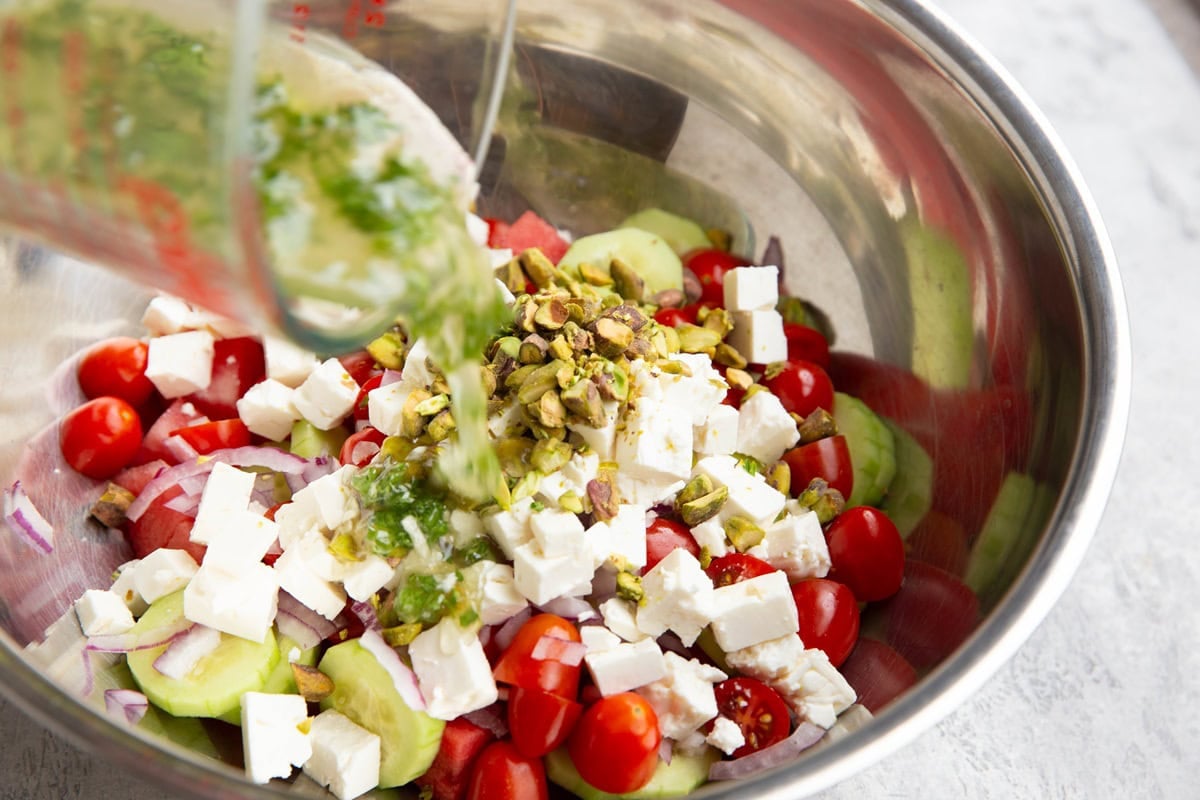 Pouring lemon parsley dressing into a bowl with watermelon salad ingredients.