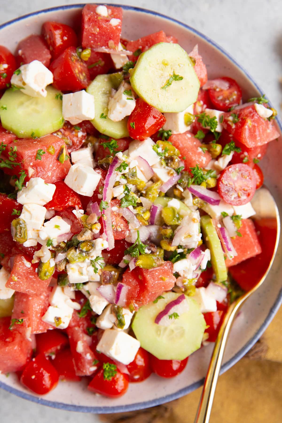 Watermelon feta salad in a large white bowl rimmed with blue with cucumbers, feta cheese, pistachios, red onions, and more. A golden spoon to the side for serving.