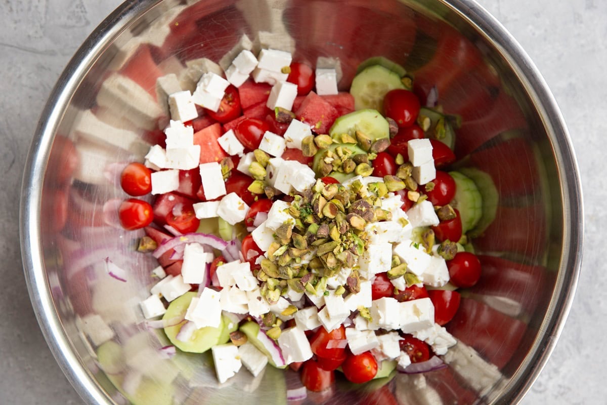 Stainless steel bowl full of chopped watermelon, cucumbers, tomatoes, feta cheese, red onions and pistachios.