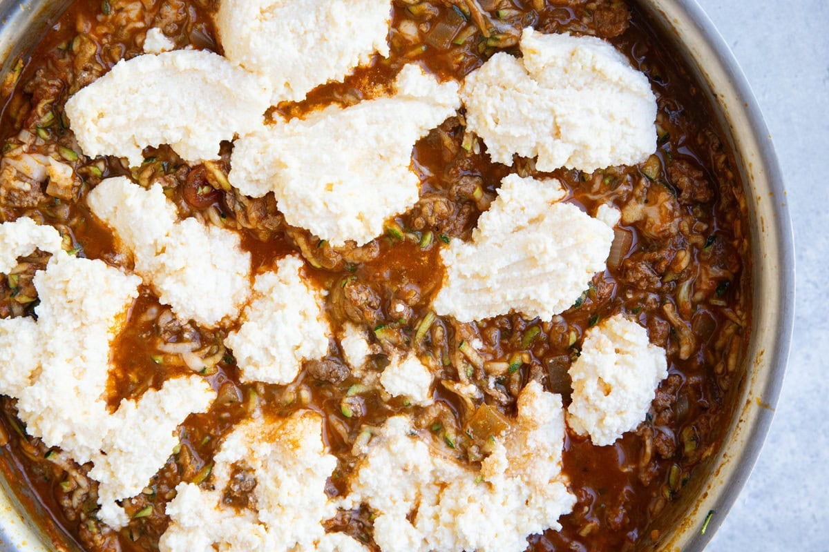 Skillet with grated zucchini mixed with meat sauce and lump of ricotta mixture added on top.