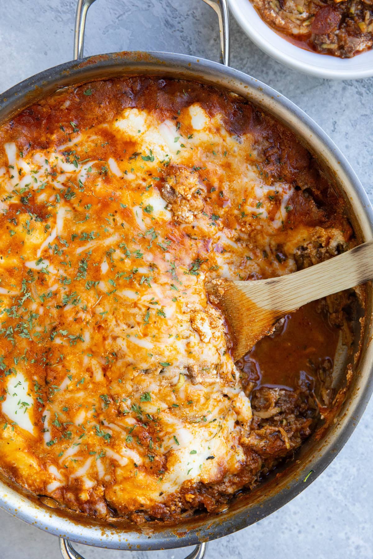 Stainless steel skillet with zucchini lasagna inside and a wooden spoon scooping some of it out. A bowl to the side with a portion of the lasagna.