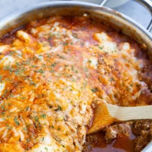 Stainless steel skillet with zucchini lasagna and a wooden spoon to scoop it out. A bowl with zucchini lasagna in the background.