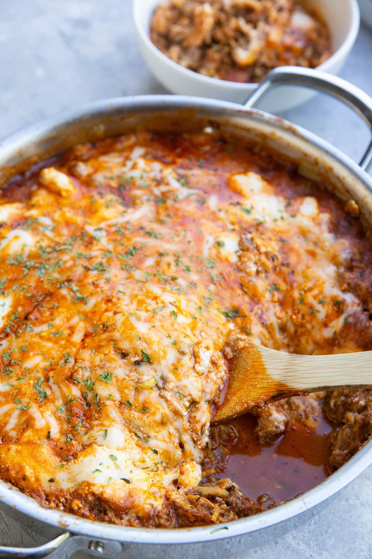 Stainless steel skillet with zucchini lasagna and a wooden spoon to scoop it out. A bowl with zucchini lasagna in the background.