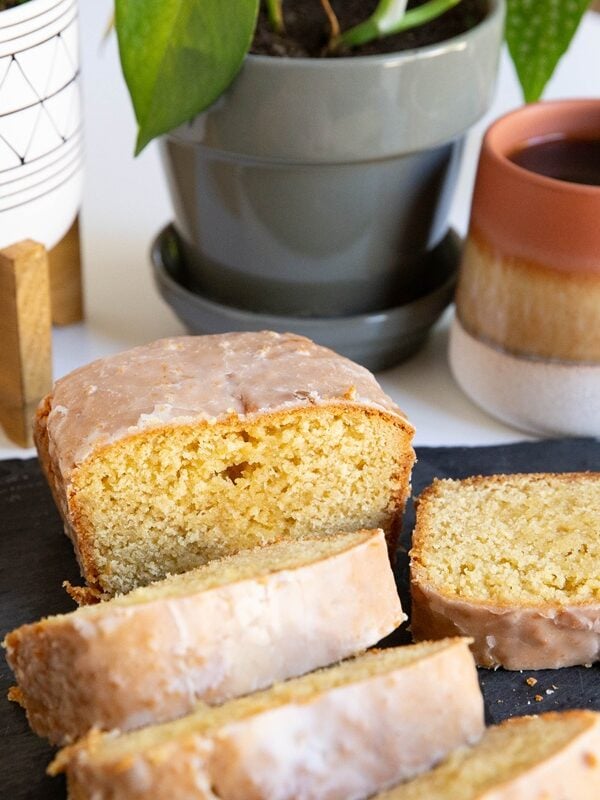 Glazed donut bread cut into slices on a black cutting board with a cup of coffee and some plants in the background.