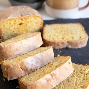 Loaf of bread with a glaze cut into thick slices on a black cutting board with a mug of coffee in the background.