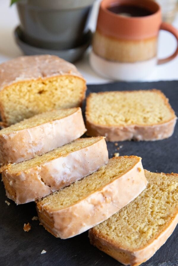 Loaf of bread with a glaze cut into thick slices on a black cutting board with a mug of coffee in the background.