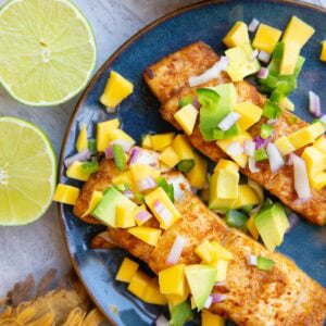 Two cooked halibut filets on a blue plate with mango salsa on top and a bowl of mango salsa to the side with a lime cut in half.