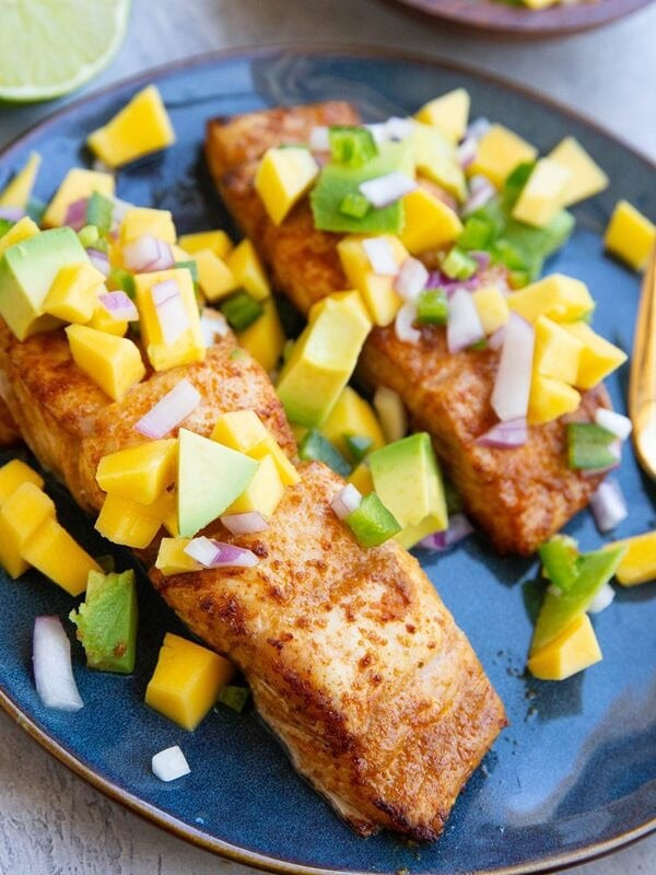 Two halibut fillets on a blue plate with mango salsa on top and a golden fork, ready to serve. A lime sliced in half sits in the background.