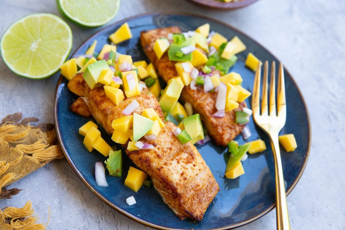 Two halibut fillets on a blue plate with mango salsa on top and a golden fork, ready to serve. A lime sliced in half sits in the background.