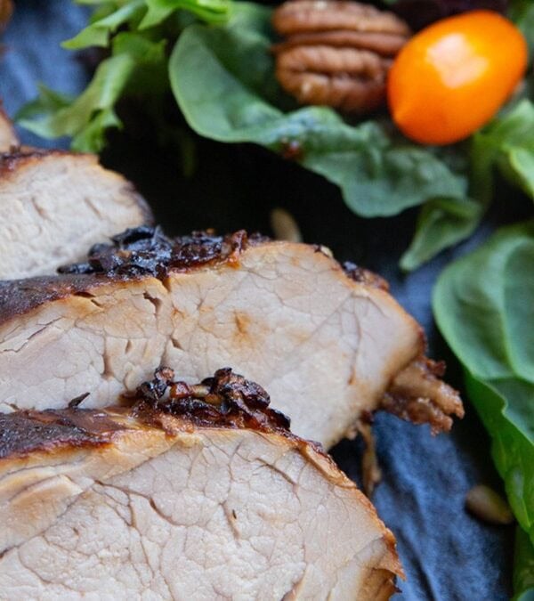 Sliced pork tenderloin on a plate with a green salad with spinach, tomatoes, dried cranberries, pecans and sunflower seeds to the side.