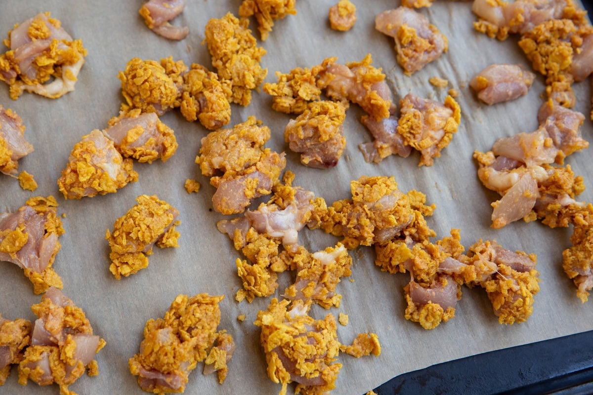 Baking sheet lined in parchment paper with raw cornflake covered chicken pieces ready to go into the oven.